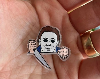 May include: A clear plastic pin with a cartoon illustration of Michael Myers from the Halloween franchise. He is holding a knife and peeking over a surface.