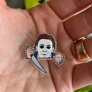 May include: A clear plastic pin with a cartoon illustration of Michael Myers from the Halloween franchise. He is holding a knife and peeking over a surface.