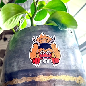 Puede incluir: Una maceta gris colgante con un borde dorado y una pegatina de un monstruo rojo con cuernos y el texto "Hellphila Club".