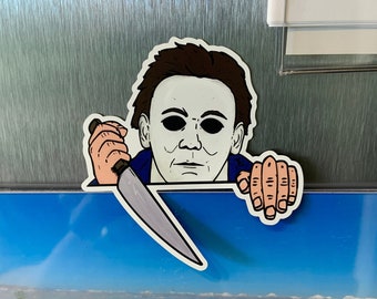 May include: A sticker of a horror movie character with a white mask and brown hair holding a knife. The sticker is on a silver refrigerator door above a photo of three dogs on a beach.