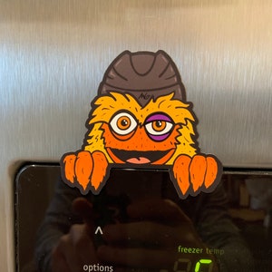 May include: A yellow and orange fuzzy monster with a black hat and a purple eye patch peeks over the top of a black refrigerator control panel. The panel displays a digital temperature reading of -08 degrees Fahrenheit.