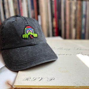May include: A black baseball cap with an embroidered monster design in green, blue and red. The monster has its tongue sticking out and is peeking out from behind the cap.