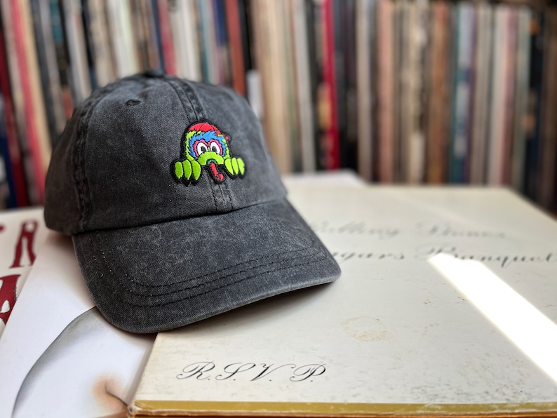 May include: A black baseball cap with an embroidered monster design in green, blue and red. The monster has its tongue sticking out and is peeking out from behind the cap.