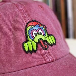 May include: A red baseball cap with an embroidered patch of a cartoon monster with a long tongue. The monster is green, blue and red.