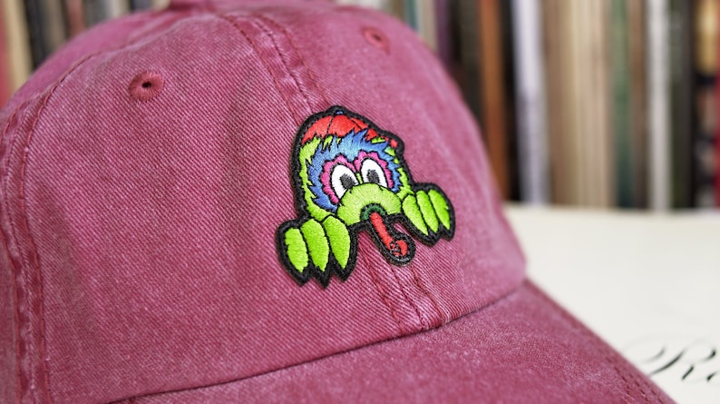May include: A red baseball cap with an embroidered patch of a cartoon monster with a long tongue. The monster is green, blue and red.