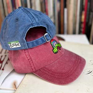 May include: Two baseball caps stacked on top of each other. The top cap is a dark blue denim with a green label that says "good for boosie". The bottom cap is a maroon colour with a green and yellow cartoon character embroidered on the front.