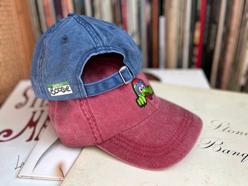 May include: Two baseball caps stacked on top of each other. The top cap is a dark blue denim with a green label that says "good for boosie". The bottom cap is a maroon colour with a green and yellow cartoon character embroidered on the front.