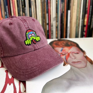 May include: A burgundy baseball cap with an embroidered green, blue and yellow cartoon monster on the front.