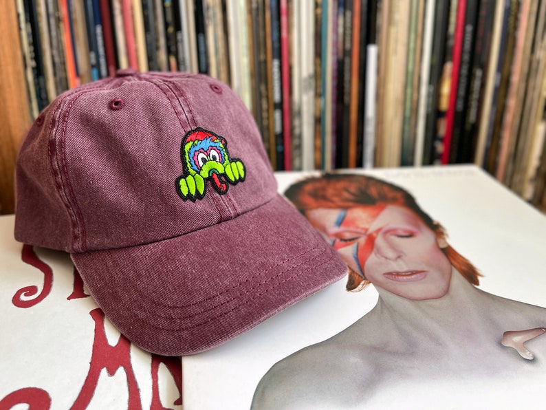 May include: A burgundy baseball cap with an embroidered green, blue and yellow cartoon monster on the front.