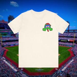 May include: A white t-shirt with a green, blue, and red cartoon monster peeking out from behind the shirt. The monster is wearing a red hat.