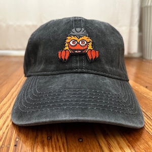 Pu&ograve; includere: Un cappello da baseball in cotone nero, usurato, con una toppa ricamata di un mostro rosso, arancione e giallo con un casco in testa.