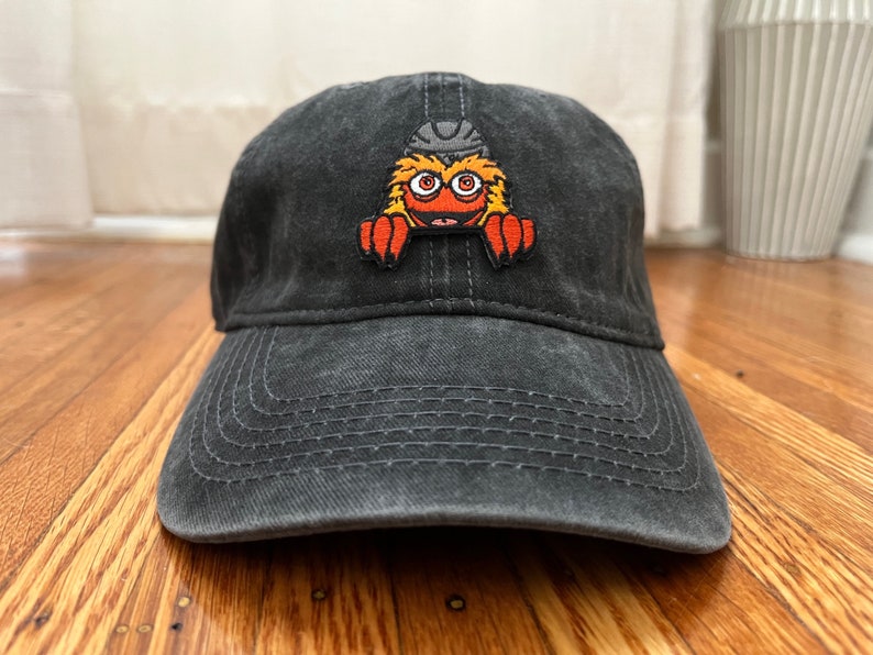 Pu&ograve; includere: Un cappello da baseball in cotone nero, usurato, con una toppa ricamata di un mostro rosso, arancione e giallo con un casco in testa.