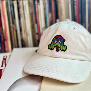 May include: White baseball cap with a green, blue and red cartoon monster patch on the front.