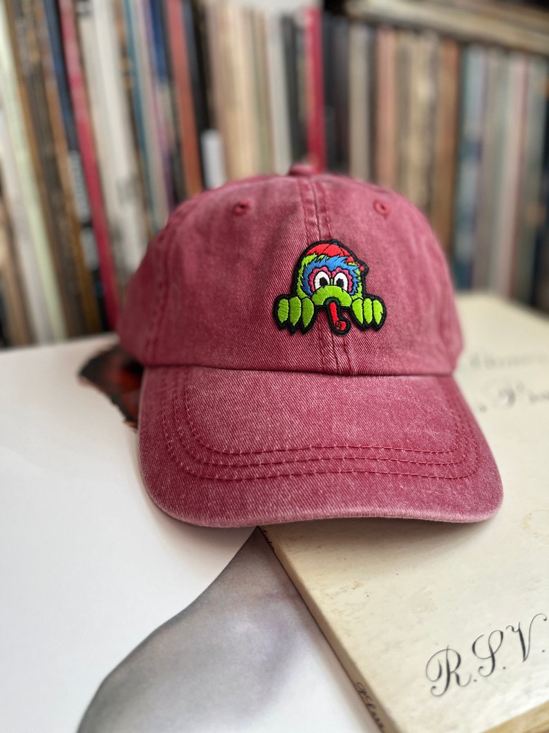 May include: A red, distressed, cotton baseball cap with a colourful cartoon monster patch on the front. The monster is green, blue, red, and yellow with a black outline. The monster has a big, toothy grin and is wearing a red hat.