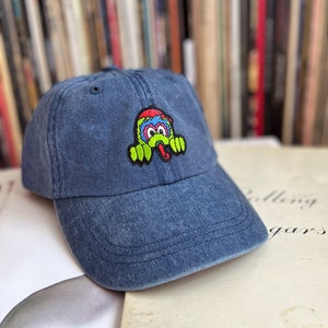 May include: A blue denim baseball cap with a colourful cartoon monster patch on the front. The monster has green claws, a red tongue, and blue and yellow eyes.