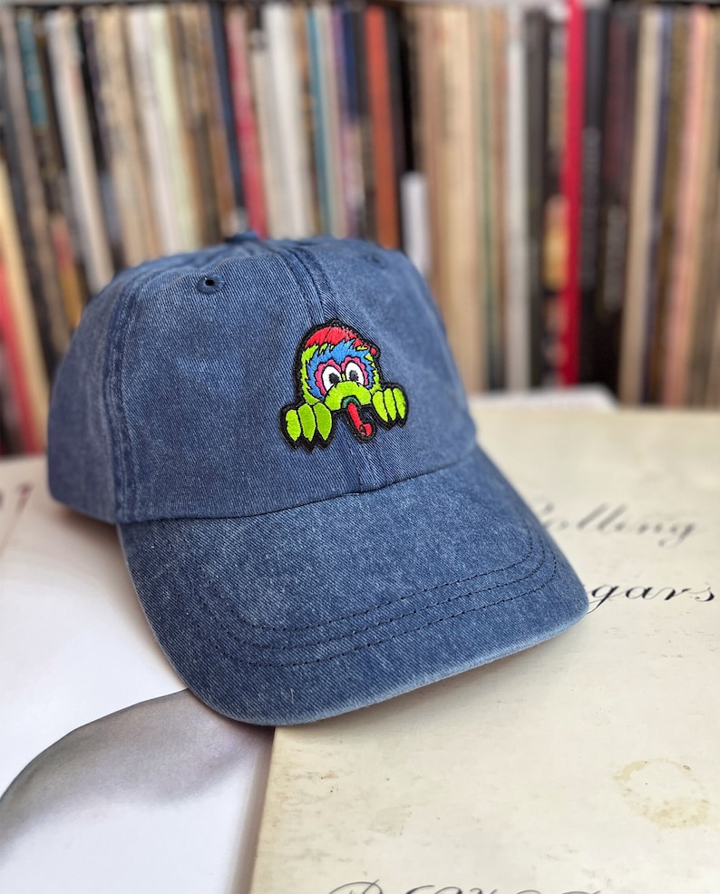 May include: A blue denim baseball cap with a colourful cartoon monster patch on the front. The monster has green claws, a red tongue, and blue and yellow eyes.