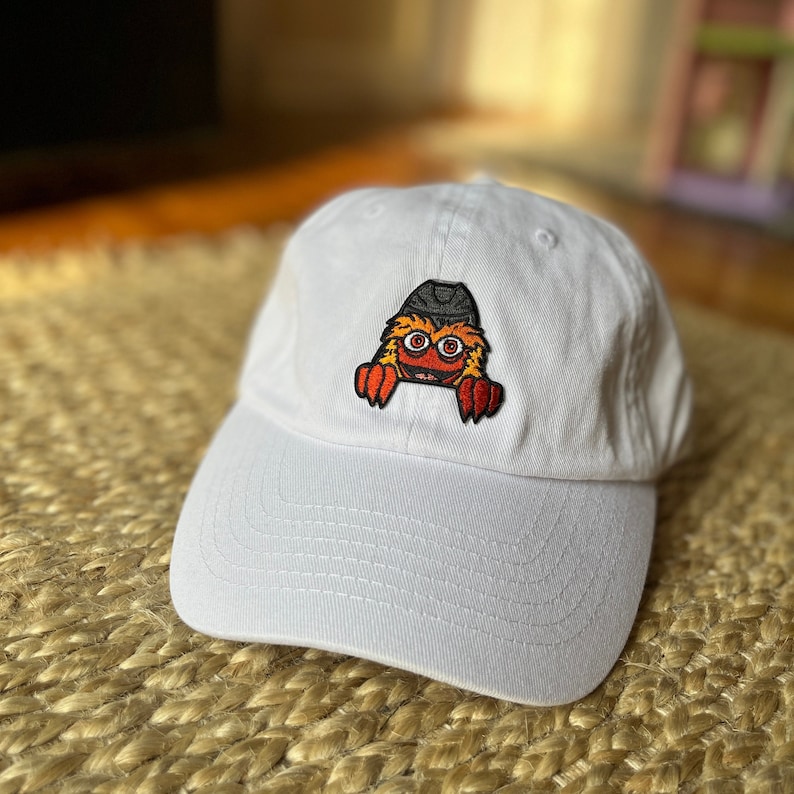 Pu&ograve; includere: Cappellino da baseball bianco con un personaggio dei cartoni animati ricamato che indossa un cappello nero. Il personaggio ha la pelliccia arancione, gli occhi rossi e una bocca bianca.