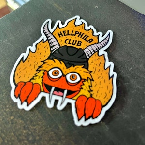 Puede incluir: Una pegatina con un monstruo de dibujos animados con pelaje naranja, ojos grandes y una sonrisa. El monstruo lleva un sombrero negro y tiene cuernos. El texto "HELLPHILA CLUB" está encima del monstruo.