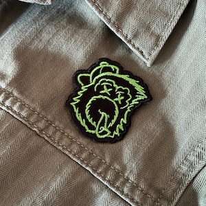 May include: Close-up of a green shirt with a black and green embroidered patch. The patch features a cartoon gorilla design with a neon green outline. The shirt has a herringbone pattern and a buttoned pocket.
