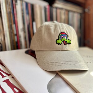 May include: A beige baseball cap with a colourful cartoon monster patch on the front. The monster has a green body, red and blue hair, and a long tongue.