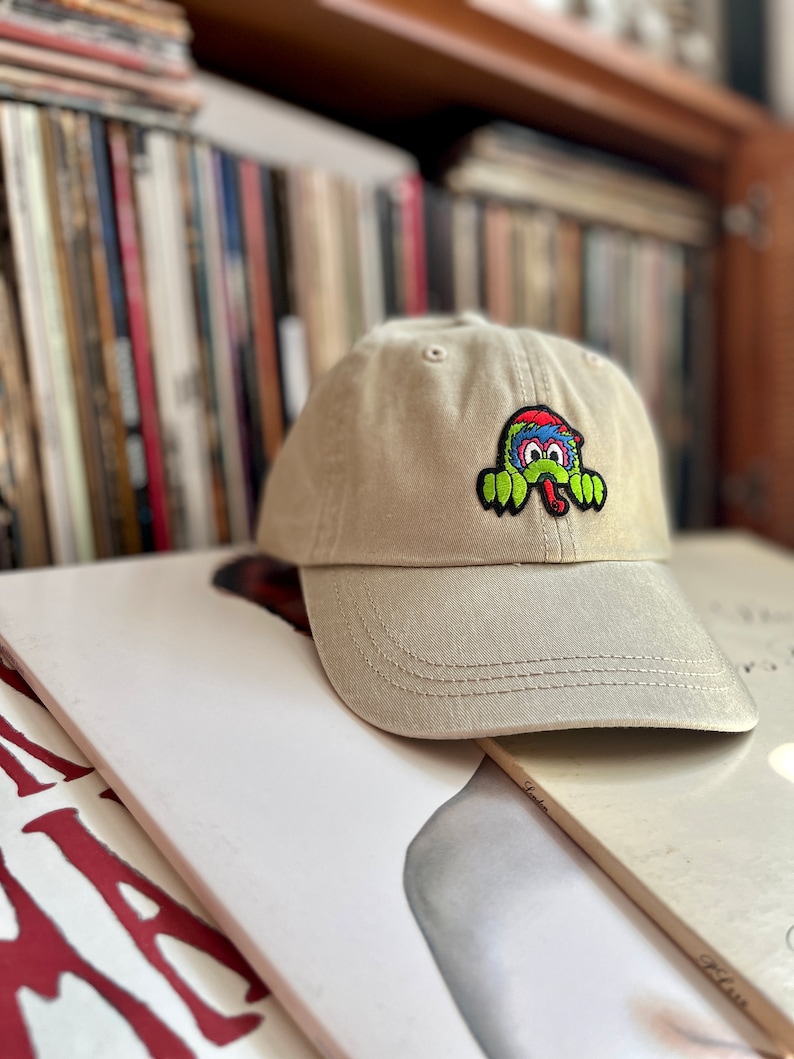 May include: A beige baseball cap with a colourful cartoon monster patch on the front. The monster has a green body, red and blue hair, and a long tongue.