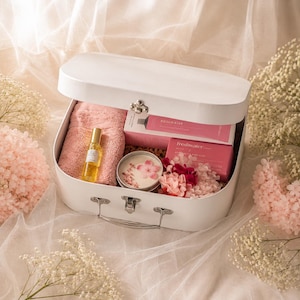 May include: A white suitcase-style gift box containing a pink towel, a small glass bottle with a gold cap, a tin candle with floral accents, and two pink rectangular boxes labelled Rosewater and Freshwater with pink and white dried flowers.
