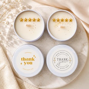 May include: Two round white metal candle tins with cream-coloured wax. Each candle features five small gold star-shaped embeds and a wooden wick. The lids are white with labels reading "thank you" and "MADE WITH LOVE THANK YOU".