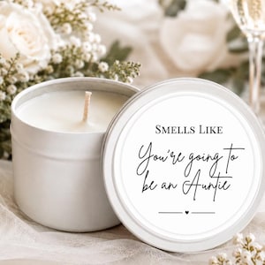 May include: A white metal candle tin with a single wick and a removable lid. The lid features a circular white label with black text that reads: SMELLS LIKE You're going to be an Auntie, with a small heart icon centred below the text.