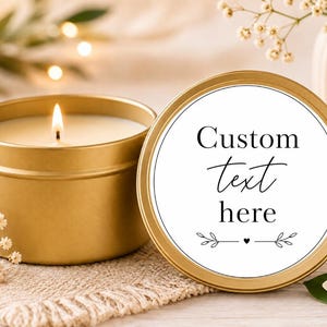 May include: A gold-coloured metal candle tin with a lit white candle inside, accompanied by a matching gold lid featuring a white circular label with the black text Custom text here and a small heart and leaf graphic design element.