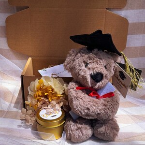 May include: A graduation gift set with a teddy bear in a black mortarboard and red bow tie. The set includes a gold candle, dried flowers, and a card, presented in a brown box. The candle is in a gold tin.