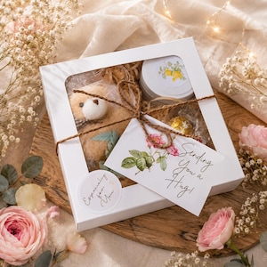 May include: A white square gift box with a clear window containing a small tan plush bear, a round metal tin with a yellow floral label, and a gold-wrapped chocolate. A tag tied with brown twine reads Sending You a Hug and Especially for You.