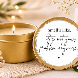 May include: A gold-coloured metal candle tin with a lit white wax candle inside. The lid is placed next to the tin, featuring a white circular label with black text that reads, "Smell's Like, It's not your problem anymore!" with a small heart.