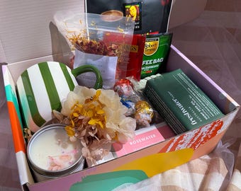 Puede incluir: Una caja de regalo llena de una taza a rayas verdes y blancas, una vela, bolsitas de té y otros artículos. La caja tiene un diseño colorido en el lateral y contiene una variedad de golosinas y productos de cuidado personal.