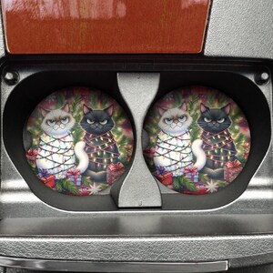 May include: Two circular car cup holder coasters featuring a festive illustration of a white cat and a black cat tangled in colorful Christmas lights. The cats are positioned in front of a pine tree with wrapped gifts at their feet.