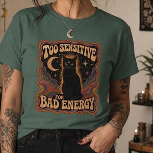 May include: A sage green short-sleeve t-shirt featuring a graphic print of a black cat sitting under a crescent moon with the text "TOO SENSITIVE FOR BAD ENERGY" in a wavy, retro-style font surrounded by colorful, swirling abstract patterns.