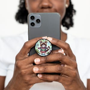 May include: A round phone grip featuring a grumpy cat wearing a Christmas sweater and reindeer antlers. The cat is surrounded by winter foliage. The phone grip is attached to a black smartphone.