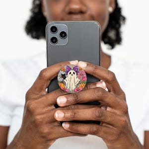 May include: A circular phone grip with a Halloween design. It features a white ghost, a black cat, pumpkins, flowers, and a purple bow. The grip is attached to a black smartphone.