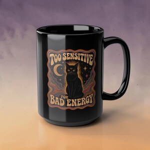 May include: A black ceramic mug featuring a graphic of a black cat with a crescent moon and stars. The text reads TOO SENSITIVE FOR BAD ENERGY in a wavy font. The design includes colorful, swirling cloud-like patterns in orange, purple, and blue.