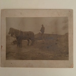 May include: A black and white photograph of a person in a hat standing next to a horse-drawn cart.