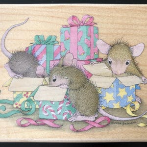 May include: A wooden stamp featuring three mice peeking out of gift boxes. The gifts are wrapped in pink, green, and blue paper with ribbons. The mice are light brown with pink ears and tails. The stamp has a festive, holiday theme.