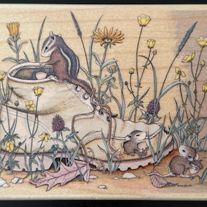 May include: A whimsical illustration of a tan work boot surrounded by wildflowers and tall grass. A chipmunk sits in the boot, while two mice are near the boot. The artwork is on a wooden block.