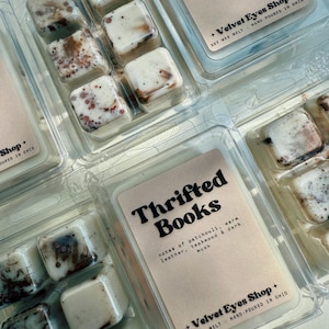 May include: Clear plastic clamshell packaging containing soy wax melts. Each package has six square melts with a marbled appearance in shades of brown and white. The label reads "Thrifted Books" with scent notes.
