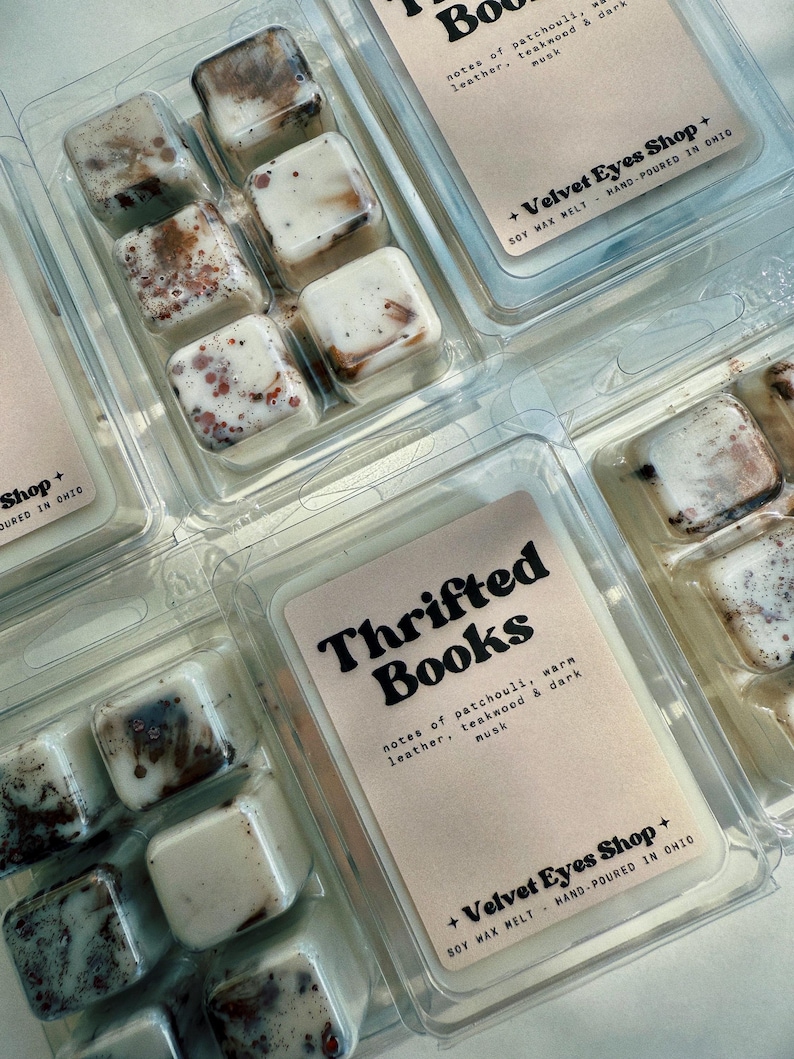 May include: Clear plastic clamshell packaging containing soy wax melts. Each package has six square melts with a marbled appearance in shades of brown and white. The label reads "Thrifted Books" with scent notes.