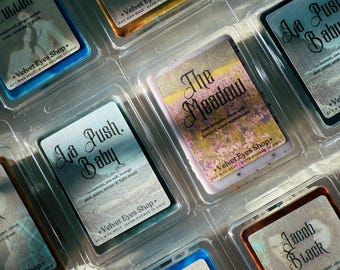 May include: A collection of scented soy wax melts in clear plastic packaging. Each melt has a label with a unique name and scent description, such as "Jacob Black" and "The Meadow". The labels feature elegant fonts and imagery.