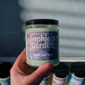 K&ouml;nnte beinhalten: Ein gr&uuml;n get&ouml;ntes Glas-Kerzenglas mit schwarzem Deckel, beschriftet mit "Sophie's Garden" mit dem Text "Velvet Eyes Shop". Das Etikett listet D&uuml;fte wie Birne, Jasmin, Rose, Flieder und Vanille auf. Weitere Kerzen sind im Hintergrund sichtbar.