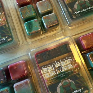 K&ouml;nnte beinhalten: Duftwachs-W&uuml;rfel in klarer Kunststoffverpackung. Die W&uuml;rfel sind in verschiedenen Farben gehalten, darunter Rot, Gr&uuml;n und Gold. Die Verpackung tr&auml;gt die Aufschrift "Sophie's Garden" mit Duftnoten und "Velvet Eyes Shop".