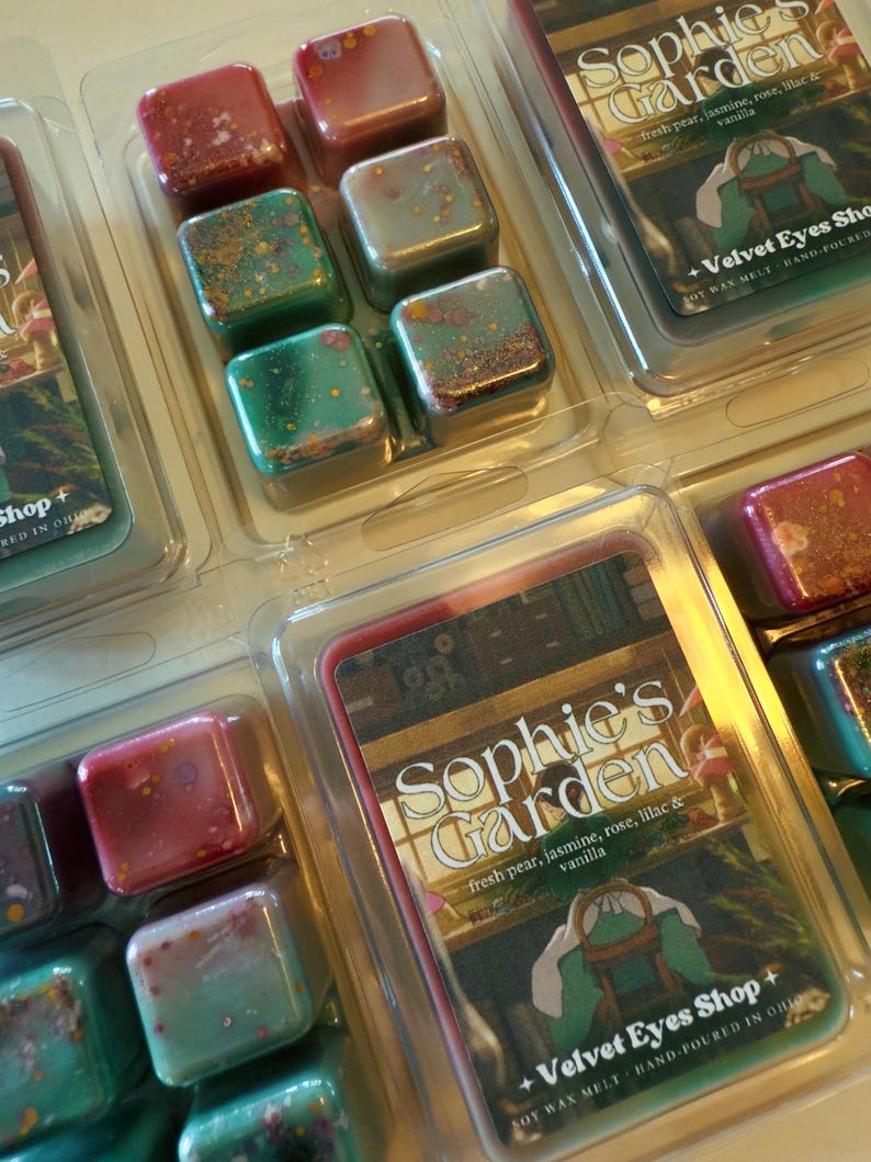 K&ouml;nnte beinhalten: Duftwachs-W&uuml;rfel in klarer Kunststoffverpackung. Die W&uuml;rfel sind in verschiedenen Farben gehalten, darunter Rot, Gr&uuml;n und Gold. Die Verpackung tr&auml;gt die Aufschrift "Sophie's Garden" mit Duftnoten und "Velvet Eyes Shop".