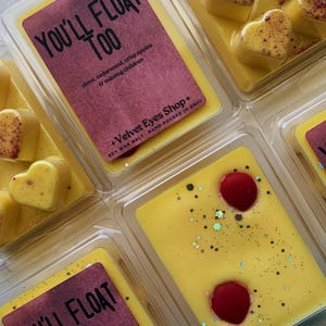 May include: Four wax melts in clear plastic containers. The wax melts are yellow with red hearts and glitter. The containers have labels that say "You'll Float Too" with a description of the scent.