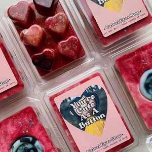May include: A set of six soy wax melts in heart and round shapes. The melts are red with glitter and feature a button design. The packaging includes the text "You're Cute As A Button" and "Velvet Eyes Shop".