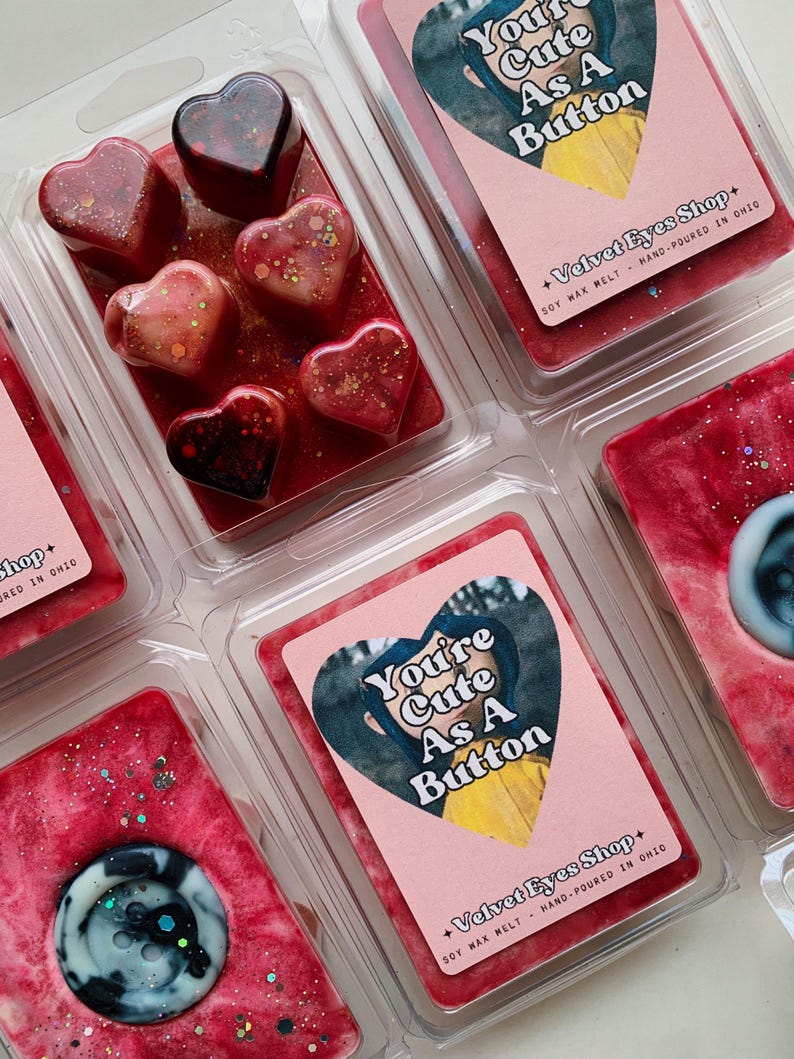 May include: A set of six soy wax melts in heart and round shapes. The melts are red with glitter and feature a button design. The packaging includes the text "You're Cute As A Button" and "Velvet Eyes Shop".
